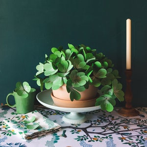 Peut inclure: Une plante de trèfle en papier vert dans un pot en terre cuite sur un support à gâteau blanc avec une tasse verte et une nappe blanche à motif floral.