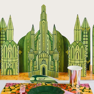 May include: A whimsical table setting featuring a green and yellow cityscape backdrop. The scene includes a floral placemat, a green plate, a pink fork, and a metallic cup. A name card reads "Garet".