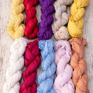 Pode incluir: Uma variedade de tiras de tecido coloridas, torcidas e dispostas em fileiras. As cores incluem amarelo, vermelho, roxo, bege, branco, rosa, azul e pêssego. O tecido parece macio e fluido, com uma superfície texturizada. O fundo é uma superfície de madeira clara e envelhecida.