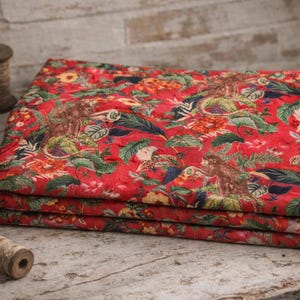 Può includere: Un tessuto rosso piegato con una stampa floreale e animale. Il tessuto è impilato su una superficie di legno, con forbici, filo e altri accessori per cucire nelle vicinanze. La stampa include foglie verdi, fiori gialli e animali marroni.