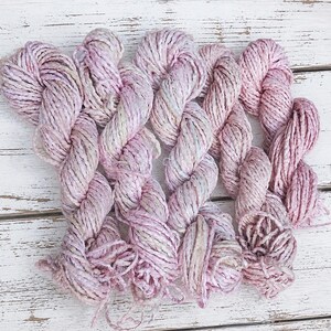 May include: Several skeins of hand-dyed yarn in shades of pink, lavender, and cream. The yarn has a textured, slightly bulky appearance, with subtle variations in color throughout. The skeins are arranged on a white wooden surface.