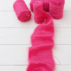 May include: Bright pink silk ribbon and spools of ribbon on a white wooden surface. The ribbon is frayed along the edges, and the spools are tightly wound. The image showcases the texture and color of the ribbon, suitable for crafts and decorative projects.