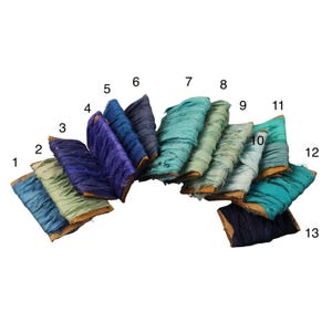 Pode incluir: Uma coleção de treze rolos de fita de seda colorida, dispostos em semicírculo. As fitas variam em tons de azul, verde e turquesa. Cada rolo é numerado de 1 a 13.