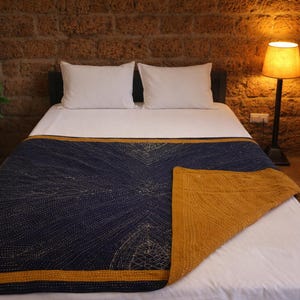 May include: A bed with white bedding, featuring a navy blue and gold quilt with a geometric pattern. The quilt has a gold border. A potted plant and a lamp are visible in the background.