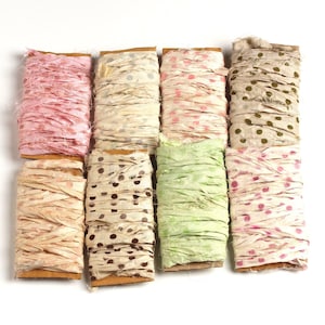 May include: A collection of eight spools of decorative ribbon in various colors and patterns. The ribbons include solid pink, cream, and green, as well as ribbons with polka dots in shades of pink, brown, and green. The spools are mounted on cardboard.