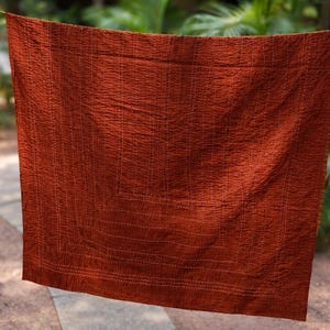 May include: A rust-colored, textured quilt hanging outdoors. The quilt is rectangular with a stitched pattern, and is suspended from a clothesline. The background includes greenery and a walkway.
