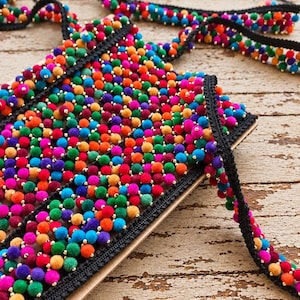 Op de afbeelding: Een decoratieve rand met een dichte schikking van kleine, veelkleurige pompons in tinten rood, oranje, geel, groen, blauw en paars. De pompons zijn bevestigd op een zwarte geweven stoffen basis, afgewisseld met kleine goudkleurige kraaltjes.