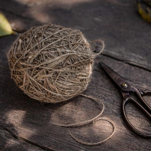 May include: A ball of twine and a pair of vintage scissors rest on a weathered wooden surface. The twine is a natural brown color, and the scissors have a dark, aged appearance. A few leaves are scattered around the twine and scissors.