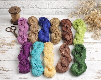 Peut inclure: Une collection d'écheveaux de fil colorés dans des tons de violet, bleu, marron, vert et jaune, disposés sur une surface en bois blanc. Une bobine en bois et de petits ciseaux sont également présents, suggérant un thème artisanal.
