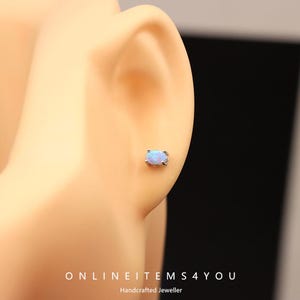 May include: A small, oval-shaped opal stud earring with a silver setting is shown in the ear. The opal has a light blue color. The text "ONLINEITEMS4YOU" and "Handcrafted Jeweller" are at the bottom of the image.
