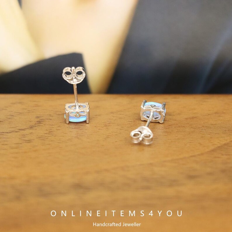 May include: A pair of silver stud earrings with square, light blue gemstones. The earrings are displayed on a wooden surface. The text "ONLINEITEMS4YOU Handcrafted Jeweller" is at the bottom of the image.
