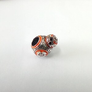 May include: A detailed, silver-toned charm featuring a BB-8 droid design. The charm has orange and black enamel accents, with intricate detailing on the spherical body and head. The design is reminiscent of the Star Wars franchise.