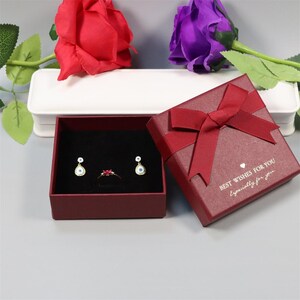 May include: A maroon gift box with a matching ribbon bow, open to reveal a pair of gold earrings and a ring. The earrings and ring have blue and red gemstones. The box has the text "BEST WISHES FOR YOU Especially for you."