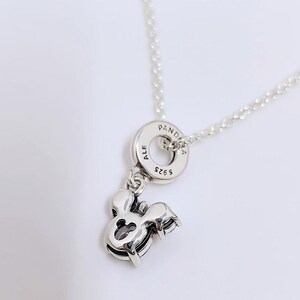 May include: A silver necklace featuring a Mickey Mouse charm. The charm is detailed with a cutout design and attached to a chain. The necklace includes a round pendant with the word "PANDORA" and the numbers "5925" engraved on it.