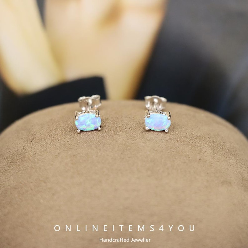 May include: A pair of oval-shaped opal stud earrings. The opals have a light blue color with hints of green and are set in silver. The earrings are displayed on a brown surface. The text "ONLINEITEMS4YOU Handcrafted Jeweller" is at the bottom.