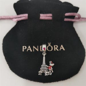 May include: A silver charm with a red heart and Eiffel Tower design hangs from a black pouch with the word "PANDORA" printed on it. The charm also features a small figure with a red hat and scarf.