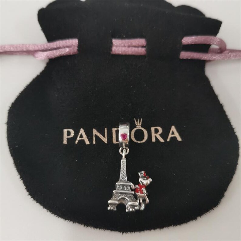 May include: A silver charm with a red heart and Eiffel Tower design hangs from a black pouch with the word "PANDORA" printed on it. The charm also features a small figure with a red hat and scarf.