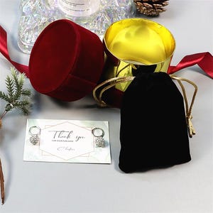 May include: A red velvet jewelry box with a gold interior sits open next to a black velvet drawstring bag. A card with the words "Thank you" and a pair of earrings are also present. A red ribbon and decorative elements complete the scene.