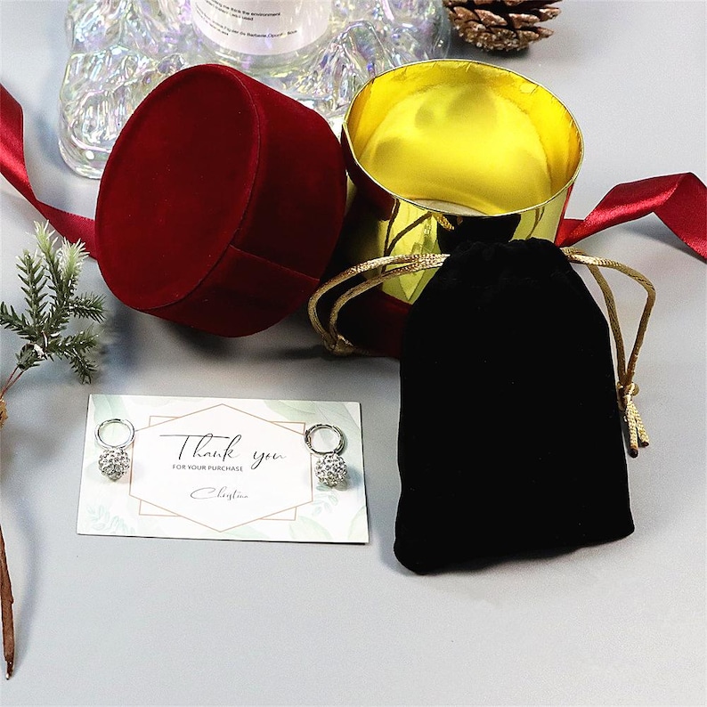 May include: A red velvet jewelry box with a gold interior sits open next to a black velvet drawstring bag. A card with the words "Thank you" and a pair of earrings are also present. A red ribbon and decorative elements complete the scene.