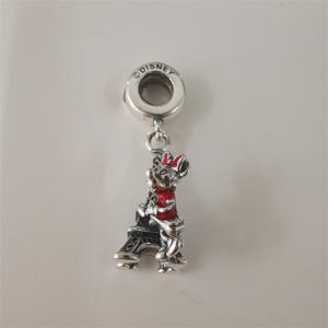 May include: A silver charm with a Disney logo at the top, featuring Minnie Mouse in a red dress and bow hugging a black Eiffel Tower. The charm is designed to be a pendant or bead.