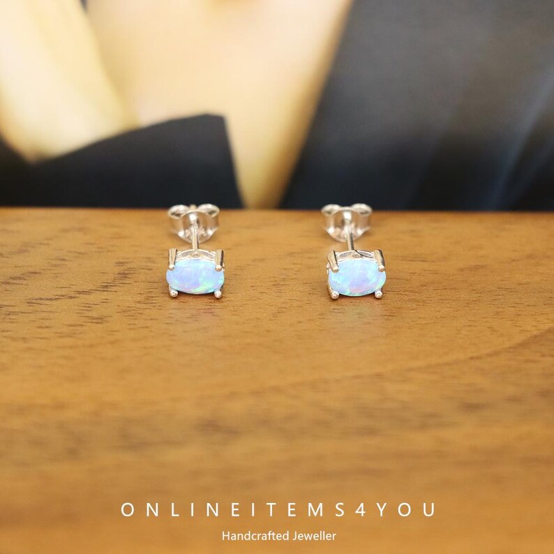 May include: A pair of stud earrings with oval-shaped, iridescent opal stones set in silver. The earrings are displayed on a wooden surface. The text "ONLINEITEMS4YOU Handcrafted Jeweller" is visible at the bottom of the image.