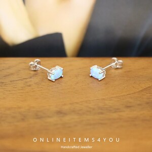 May include: A pair of silver stud earrings with oval, light blue opal stones. The earrings are set in a silver bezel setting and are displayed on a wooden surface. The text "ONLINEITEMS4YOU Handcrafted Jeweller" is visible.