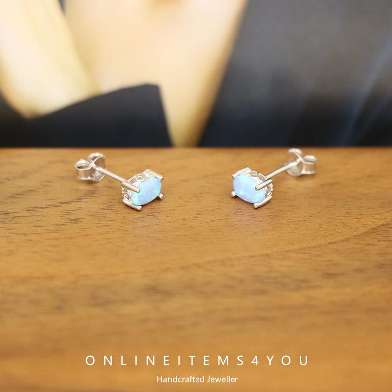 May include: A pair of silver stud earrings with oval, light blue opal stones. The earrings are set in a silver bezel setting and are displayed on a wooden surface. The text "ONLINEITEMS4YOU Handcrafted Jeweller" is visible.