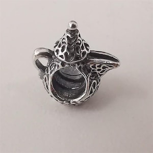 Puede incluir: Un charm plateado detallado con forma de tetera. El charm presenta intrincados patrones calados en el cuerpo, la tapa y el pico. El asa y el pico están claramente definidos, y una pequeña bola se encuentra sobre la tapa.