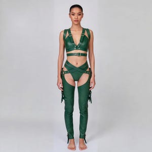 May include: A green leather harness set featuring a top with straps and a matching bottom with crisscross detailing. The set includes chaps with buckle and strap accents. The model is standing in front of a plain background.
