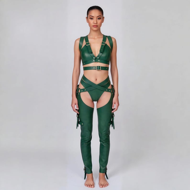 May include: A green leather harness set featuring a top with straps and a matching bottom with crisscross detailing. The set includes chaps with buckle and strap accents. The model is standing in front of a plain background.