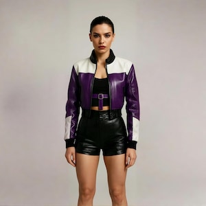 Puede incluir: Chaqueta de cuero corta con bloques de color morado, blanco y negro. La chaqueta tiene cuello alto y cremallera. La modelo lleva shorts negros y un top negro con cinturón morado.