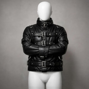Puede incluir: Chaqueta de cuero negro con múltiples correas y hebillas, exhibida en un maniquí. La chaqueta presenta un cuello alto y está diseñada con varias correas en el pecho y los brazos, aseguradas con hebillas plateadas.