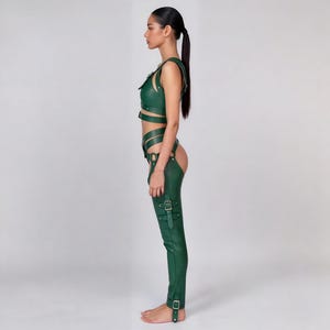 May include: A green leather outfit featuring a crop top with straps and buckle details, paired with matching chaps. The chaps have multiple straps and buckles, and the outfit is designed with a form-fitting silhouette. The outfit is made of a smooth, shiny material.