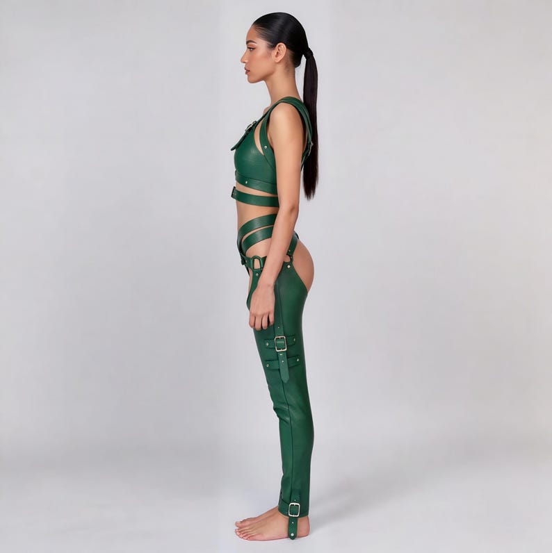 May include: A green leather outfit featuring a crop top with straps and buckle details, paired with matching chaps. The chaps have multiple straps and buckles, and the outfit is designed with a form-fitting silhouette. The outfit is made of a smooth, shiny material.