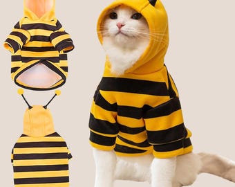 May include: A white cat wearing a yellow and black striped bee costume with a hood and antennae. The image also shows two additional bee costumes. The costumes are designed for pets.