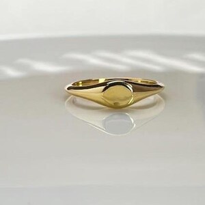 Könnte beinhalten: Ein goldener Siegelring mit poliertem Finish. Der Ring hat eine abgerundete ovale Oberfläche und einen glatten, konischen Ring. Der Ring wird auf einer weißen Oberfläche präsentiert.