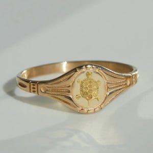 Puede incluir: Anillo dorado con diseño de tortuga. El anillo tiene una banda con grabados decorativos y un óvalo central con un emblema de tortuga dorada sobre fondo crema. El diseño es detallado y elegante.