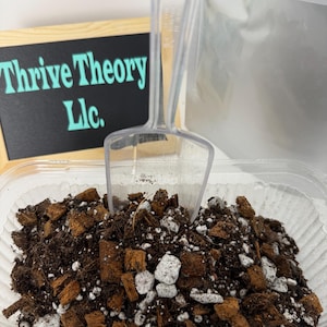 May include: A clear plastic container filled with a dark brown soil mix, including wood chips and white stones. A clear plastic scoop is inserted into the soil. A small chalkboard sign reads "Thrive Theory Llc."