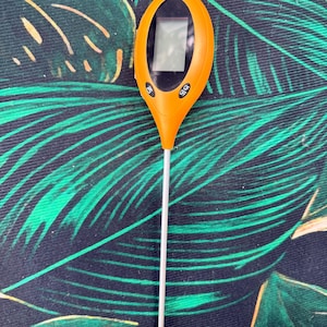 May include: An orange soil tester with a digital display and a long, silver probe. The device has buttons labeled "ON" and "功能". A red light is visible at the top. The background is a dark fabric with green leaf patterns.