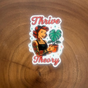 May include: A sticker featuring a woman with red hair and tattoos holding a potted plant. The sticker has the words "Thrive Theory" in a cursive font with a pink and white color scheme. The woman is wearing a black top.