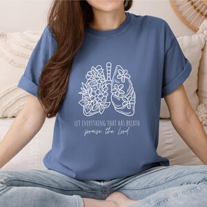 May include: A blue t-shirt with rolled-up sleeves featuring a white graphic of lungs adorned with floral designs. The text "LET EVERYTHING THAT HAS BREATH praise the Lord" is printed below the graphic. The shirt is worn by a person.