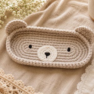 May include: A beige crochet bear-shaped tray with black eyes and nose. The tray is oval and has two small ears. The tray is on a beige fabric surface with lace trim and flowers.