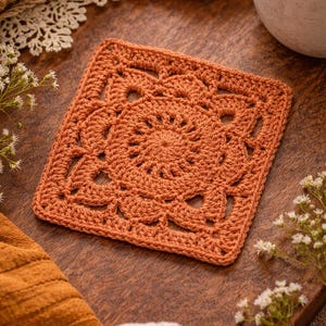 May include: A square, terracotta-colored crochet coaster with a floral design in the center. The coaster is placed on a wooden surface, with a cup and other textiles in the background. The coaster is approximately 5 inches by 5 inches.