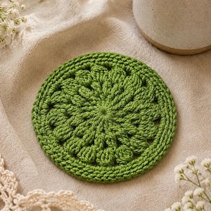 May include: A handmade, round, green crochet coaster with a floral design. The coaster is made of a thick yarn and sits on a beige linen surface. A white ceramic mug with a lemon slice is in the background.