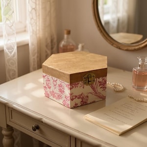 May include: A hexagonal wooden box with a light wood top and a patterned base. The base features a white background with a red floral design. The box has a brass clasp and is sitting on a white table.