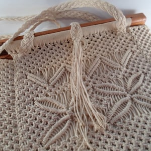 May include: A beige macrame bag with a wooden top and a long fringe. The bag is made of a thick, knotted cord and has a simple, geometric design.
