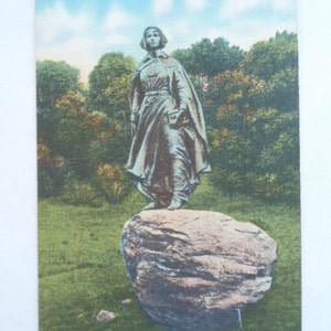 May include: Vintage postcard featuring the Pilgrim Maiden statue in Plymouth, Massachusetts. The statue is bronze-colored and stands on a large rock, set against a backdrop of trees and a blue sky. The text "Pilgrim Maiden, Plymouth, Mass." is printed at the top.