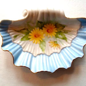May include: A decorative, shell-shaped dish with a scalloped edge. The dish features a light blue border and a white interior with a hand-painted floral design of yellow daisies and green leaves. The rim is trimmed with gold.