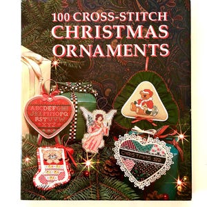 May include: A book titled "100 Cross-Stitch Christmas Ornaments" is displayed, surrounded by various cross-stitched ornaments. The ornaments include a heart, an angel, a Christmas tree, and a stocking, all with festive designs and colors.