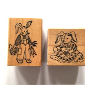 May include: Two wooden stamps with bunny illustrations. One stamp features a bunny in overalls holding a basket and carrots. The other stamp shows a bunny playing an instrument. Both stamps have a light wood grain.